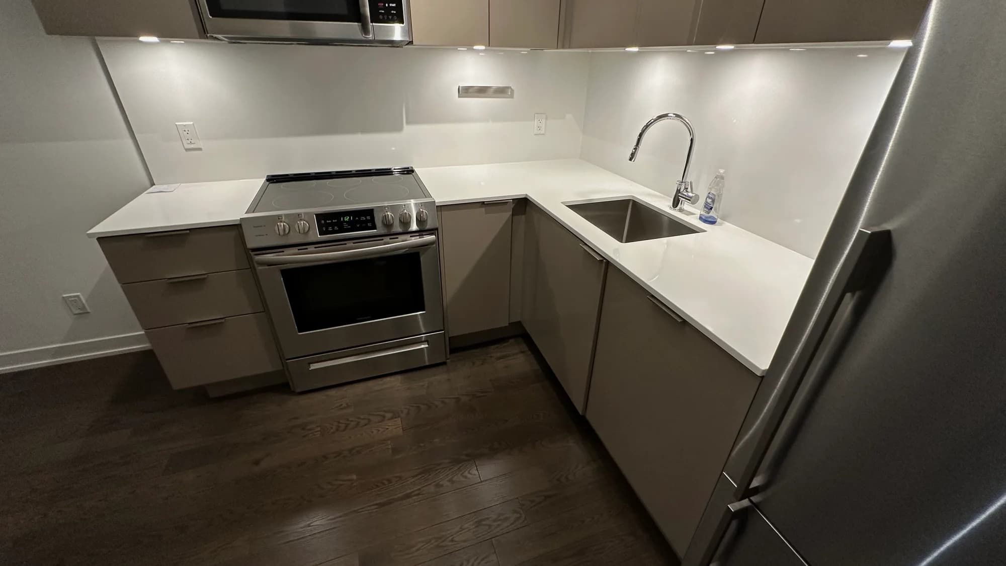 Compact L-shaped kitchen with taupe cabinetry, white backsplash, and polished counters after cleaning